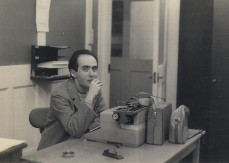 Foto em preto e branco de um homem, branco, de cabelos curtos, lisos e escuros, sentado em uma mesa com uma máquina de escrever à sua frente. Ele veste blazer e camiseta, tem os cotovelos apoiados sobre a mesa e uma das mãos próxima do rosto. Na mesa, há um cinzeiro, um grampeador e bolsas. Atrás dele, uma parede clara e uma porta aberta