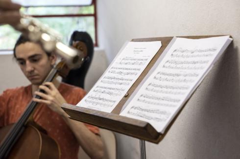 Foto com foco em  uma estante de partituras com duas folhas de partitura. Ao fundo há um homem desfocado tocando violoncelo. Ele é branco, tem cabelos castanhos presos e usa camiseta vermelha. Próxima à câmera, à esquerda da foto, também desfocada, há a mão de uma pessoa tocando flauta transversal. O ambiente tem paredes brancas e uma janela com vista para árvores.