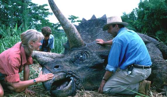 Cena do filme Jurassic Park: O Parque dos Dinossauros. Um dinossauro, triceratops, está deitado, possivelmente morto. Uma mulher e um homem estão abaixados na frente do animal e cada um tem uma de suas mãos encostada nele. Atrás do dinossauro, um menino em pé, com as mãos nos joelhos, olha para o animal. A mulher sorri, tem pele branca, cabelos loiros e veste uma camisa vermelha com as mangas dobradas. O homem tem pele branca, cabelo escuro, veste camisa azul, calça bege e usa chapéu bege. O menino tem pele branca, cabelos claros e usa roupa azul. Árvores e plantas formam o plano de fundo.
