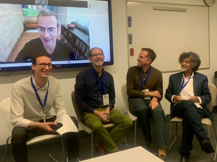 Foto de Almir Almas com outros professores na Sixteenth Global Studies Conference da OBU. Cinco homens sorriem. Quatro deles estão sentados. Um deles aparece em uma tela atrás dos outros em uma videochamada. Quatro têm pele branca, usam calças e camisas e um tem pele negra e usa calça e blazer pretos sobre a camisa branca. Os que participam presencialmente usam crachás. Dois homens à direita da imagem olham para a tela, os da esquerda olham para frente.