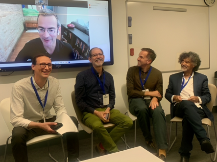 Foto de Almir Almas com outros professores na Sixteenth Global Studies Conference da OBU. Cinco homens sorriem. Quatro deles estão sentados. Um deles aparece em uma tela atrás dos outros em uma videochamada. Quatro têm pele branca, usam calças e camisas e um tem pele negra e usa calça e blazer pretos sobre a camisa branca. Os que participam presencialmente usam crachás. Dois homens à direita da imagem olham para a tela, os da esquerda olham para frente. 