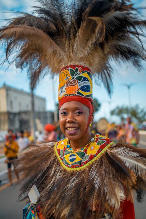 Foto de uma mulher negra usando roupas adornadas por grandes plumas no chapéu e no colar. O chapéu tem ilustrações de uma onça e da bandeira do Brasil e o colar tem ilustrações de flores e da bandeira do Brasil. A mulher sorri para a câmera.