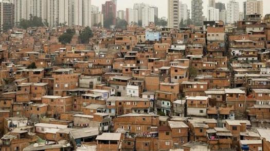 Fotografia colorida mostra a Favela de Paraisópolis vista do bairro Morumbi, em São Paulo. Em primeiro plano há várias casas de alvenaria espalhadas de maneira desordenada ao longo dos diferentes níveis do terreno. Ao fundo, diversas torres de condomínios residenciais de classe média e classe média alta.