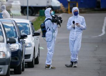 Foto colorida mostra um repórter e um cinegrafista paramentados com roupas e máscaras para evitar a contaminação pelo coronavirus.