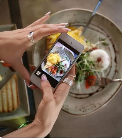 Foto a partir de cima de mãos de uma pessoa branca com anéis prateados e unhas compridas e pintadas de cor clara segurando um celular que fotografa um prato de cerâmica com alimentos de diferentes cores. No prato há talheres e ele está sobre uma superfície clara.