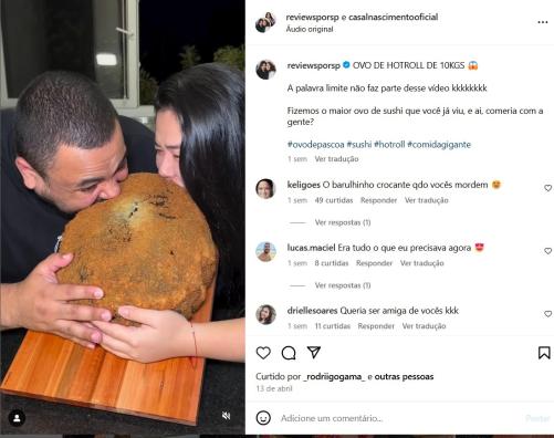 Foto de homem pardo e mulher amarela comendo um hot holl gigante em formato de ovo. O homem está à esquerda e tem cabelos curtos e escuros, a mulher está à direita e tem cabelos longos, lisos e escuros. Ambos vestem preto e têm uma das mãos segurando o hot roll, que está sobre uma tábua de madeira ao centro. No fundo da imagem, há uma janela de vidro e o céu escuro.
