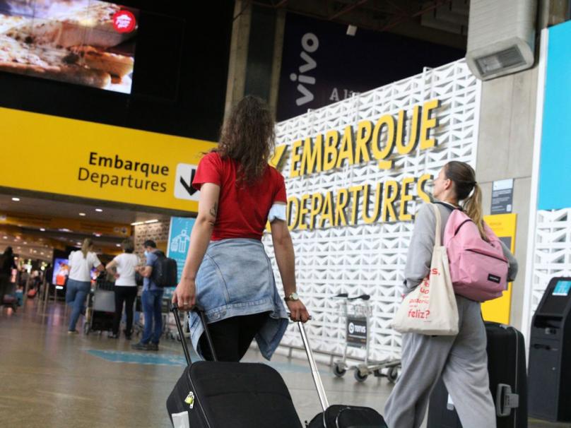 Foto de duas jovens brancas vestidas com roupas leves e caminhando com bolsas e malas em direção à área de embarque de um aeroporto. Em segundo plano, aparecem em destaque placas e estruturas com os dizeres Embarque - Departures, nas cores amarela, branca e preta. Diversas outras pessoas circulam pelo local. 