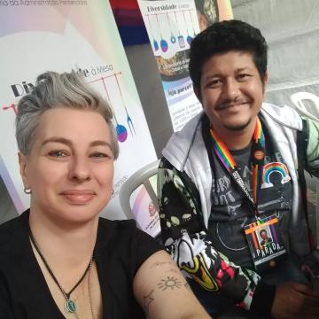 Na foto há duas pessoas. À esquerda, Paula Curi, uma mulher branca, de cabelo grisalho e curto. Sorri, veste uma camiseta preta, colares, brincos e um de seus braços expõe tatuagens. A sua direita, Elvis Stronger, um homem negro de cabelo curto e barba pretos. Sorri, usa um casaco com estampas e carrega um crachá com as cores do arco-íris onde é possível ler "DIRETORIA" e "PARADA SP", além do seu nome.