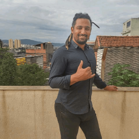 Foto de Ivan Barbosa. Homem negro, com cavanhaque e dreads no cabelo escuro, usa camisa cinza escuro de mangas dobradas e calça jeans. Ele sorri para a câmera enquanto apoia a mão esquerda em uma mureta atrás dele e faz o sinal de hang loose com a mão direita. Ao fundo, a vista do teto de algumas casas, prédios baixos e montanhas com vegetação.