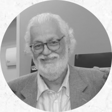 Foto em preto e branco de Edmir Perroti, um homem branco, com cabelos brancos curtos e ligeiramente ondulados e barba curta. Usa óculos com haste escura. Veste um paletó e camisa branca. 