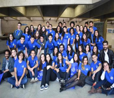 Foto de vários alunos olhando para a câmera e sorrindo. A grande maioria veste uma camiseta azul com o logo da Jornalismo Júnior (que é o nome da entidade em letras brancas).