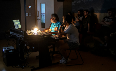 Professor e aluno estão sentados e olham para a tela de um computador. À frente deles também há uma mesa de som, iluminada por uma luminária. Ao fundo, outros alunos observam o trabalho.