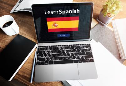 Notebook aberto com dizer &quot;Learn Spanish&quot; e bandeira da Espanha em frente à fundo preto, em cima de uma mesa de madeira com outros objetos
