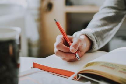 Foto colorida em ambiente interno, da mão de uma pessoa segurando uma caneta e escrevendo em um caderno.