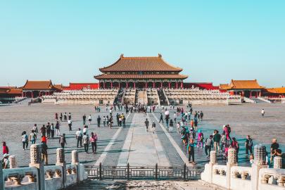 Foto colorida em ambiente externo, da Cidade Proibida na China, retratada por um pátio cheio de pessoas e uma construção de arquitetura tradicional chinesa ao fundo.