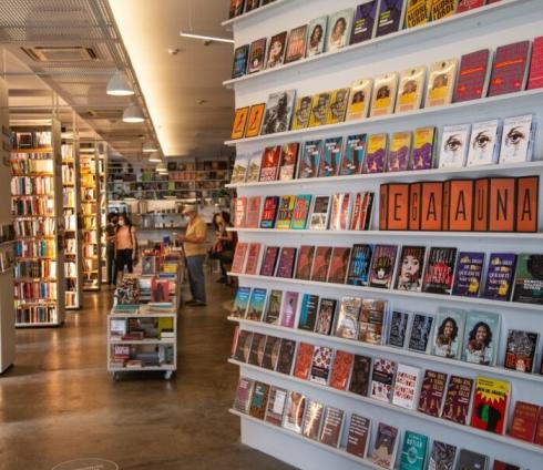 Fotografia das estantes da livraria com algumas pessoas ao fundo mexendo nos livros. No primeiro plano uma estante com diversos livros com capas nas cores vermelho, amarelo, azul e marrom.