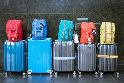 Foto colorida, em ambiente interno, de 5 malas enfileirada, com mochilas dispostas em cima