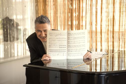 Foto de um homem branco de cabelos curtos e grisalhos, que sorri para a câmera e traja camisa branca e terno preto. Ele está sentado ao piano e apoia uma partitura com as páginas abertas sobre o instrumento.  A superfície do piano reflete a partitura e um cortinado laranja que está ao fundo. Há outras partituras sobre o instrumento.