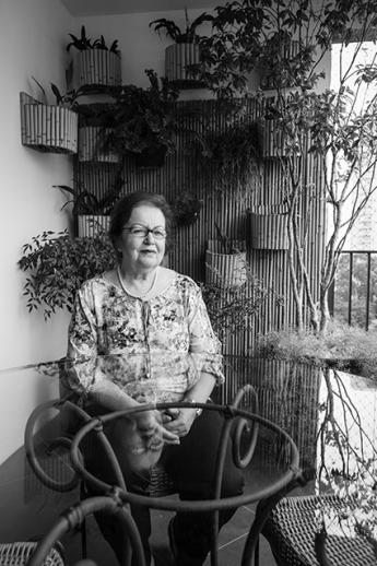 Foto em preto e branco de uma mulher idosa sentada atrás de uma mesa de vidro. Ela usa óculos, tem cabelos curtos e escuros, é branca, usa uma blusa florida. Na parede ao fundo estão penduradas algumas plantas.