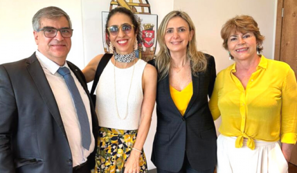 Foto de quatro pessoas posando em de pé para uma foto. À esquerda, um homem branco, de cabelos grisalhos e óculos. Ele veste uma camisa branca, uma gravata azul e um paletó cinza escuro. Ao seu lado, uma mulher branca, de cabelos escuros e ondulados, presos. Ela veste uma camisa branca, uma saia estampada nas cores amarelo, verde escuro, preto e branco, e óculos escuros. Ao seu lado outra mulher, loira de cabelos compridos. Ela veste um paletó preto e uma blusa amarela. Por fim, mais um mulher, de cabelos curtos e loiro escuros, vestindo uma camisa amarela e uma calça branca.