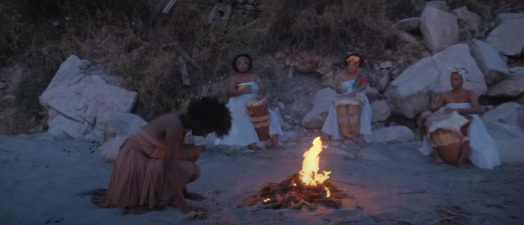 Em uma praia, três mulheres negras vestidas de branco tocam instrumentos de percussão ao redor de uma fogueira enquanto outra mulher negra, em destaque e em vestido amarelo, dança.