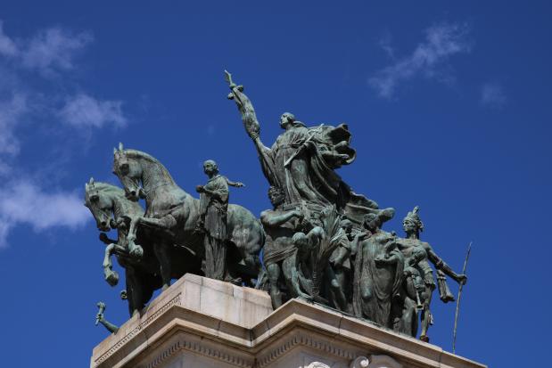 Foto de uma parte do Monumento à Independência, o conjunto escultórico Marcha Triunfal da Nação Brasileira. Dois cavalos estão à frente de um grupo de personagens: no alto, uma mulher com vestes esvoaçantes e um mastro nas mãos representa o Brasil; ao seu lado, homens e mulheres com diferentes tipos de vestimenta, incluindo um homem indígena, carregam artefatos como cruz, lança e tocha. A obra, que tem aparência desgastada, está apoiada em uma base de granito branco com adornos nas bordas. Ao fundo, o céu é bem azul e com poucas nuvens.