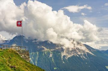 Foto colorida em ambiente externo, da bandeira da Suíça hasteada em frente a uma montanha