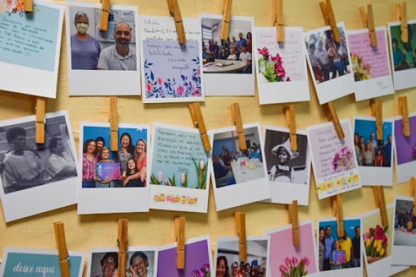 Foto de um mural de recados. O mural é de madeira clara, possui barbantes horizontais e neles estão, presas por prendedores de roupa, fotos de flores e de Silvia com colegas de trabalho com recados escritos nas margens.