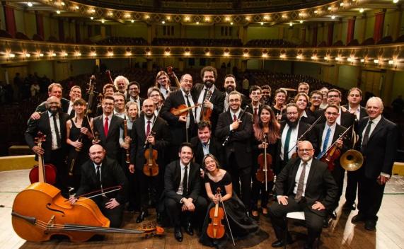 Foto dos 32 instrumentistas da Orquestra de Música Antiga posando sorridentes. São homens e mulheres de diversas idades, alguns sentados e outros de pé. Eles estão sobre um palco, com a plateia vazia ao fundo. É possível identificar diversos instrumentos, como viola, contrabaixo, violino, violoncelo e trompa.