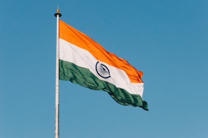 Foto colorida em ambiente externo, da bandeira da Índia balançando em frente ao céu azul