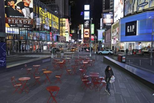 Foto da Times Square. Muitos prédios exibem diversas propagandas luminosas em telas digitais. Mesas e cadeiras que estão em primeiro plano aparecem vazias. Há algumas pessoas e alguns carros nas ruas. 