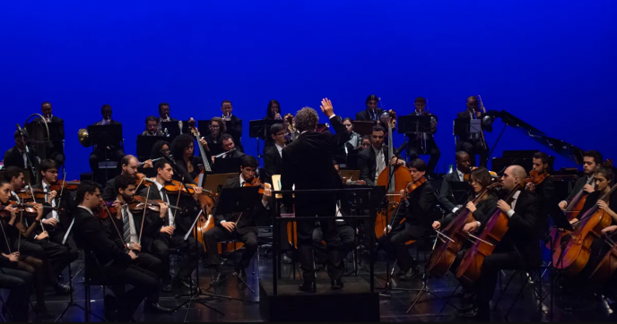 Foto de uma orquestra tocando. No centro, encontra-se o maestro em cima de um púlpito, de onde rege os músicos de costas para a câmera. Ao redor dele, os instrumentistas se aglomeram sentados, enquanto tocam. Todos vestem ternos pretos, no caso dos homens, e vestidos da mesma cor, no caso das mulheres. O fundo do palco é azul.