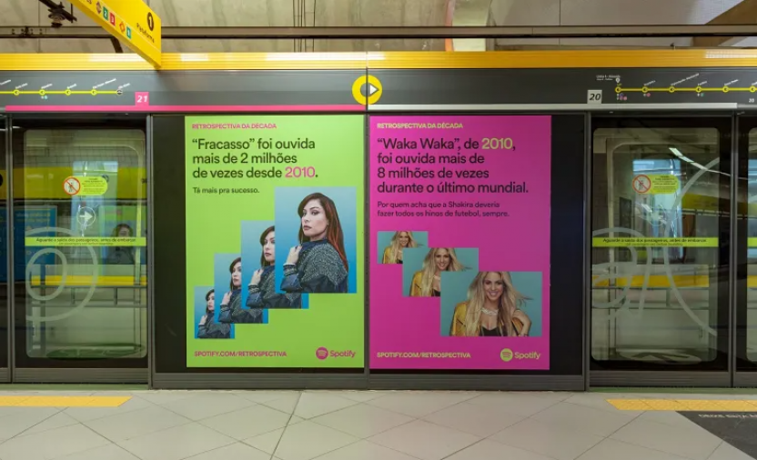 Foto das portas de vidro que separam os trilhos do metrô dos passageiros na plataforma de embarque. Em cada porta está exposta uma propaganda. À esquerda há uma arte com fundo verde-limão que cita a música “Fracasso” e exibe a mesma foto da cantora Pitty repetida 5 vezes. Pitty é branca, tem cabelos longos vermelhos e usa roupas pretas). À direita, a divulgação menciona a canção “Waka Waka”, da Shakira, com 3 fotos repetidas da artista, que é branca, tem longos cabelos loiros e veste preto e amarelo.