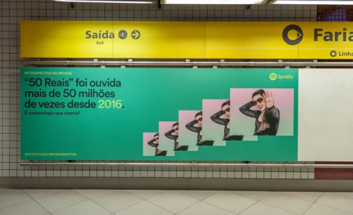 Foto de um painel exposto em um corredor de uma estação de metrô. O painel é verde e tem, à esquerda e em letras pretas, o texto “ ‘50 reais’ foi ouvida mais de 50 milhões de vezes desde 2016. É numerologia que chama?”. À direita, aparecem 5 vezes a mesma foto da cantora da música, Naiara Azevedo, que é branca, tem cabelos longos e lisos escuros, usa óculos de sol, veste uma roupa preta e usa batom vermelho. Ela sorri, coloca uma mão atrás da cabeça e com a outra acena para a câmera. O logo do Spotify aparece acima das fotos.