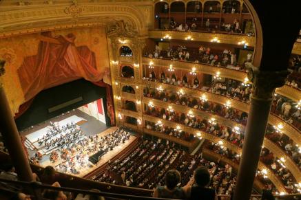 Foto de sala de concerto grandiosa e de decoração rebuscada, com seis andares de camarotes, todos lotados, bem como a plateia. No palco, está uma orquestra completa. O espaço onde as pessoas estão sentadas, assim como o palco, estão bem iluminados por uma luz amarelada.