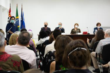 Foto da cerimônia. Em primeiro plano, estão diversas pessoas sentadas de costas. Na frente delas, à esquerda, está um homem em pé discursando. Ele usa óculos e máscara e veste roupas azuis. Atrás dele há bandeiras hasteadas. À direita, quatro pessoas estão sentadas em frente a uma mesa.