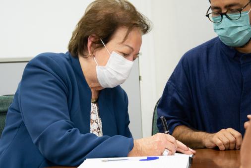 Foto de uma senhora assinando um papel apoiado em uma mesa de madeira. Ela é branca, tem cabelos curtos e lisos, veste uma blusa branca e um blazer azul e usa máscara. Ao lado dela, está um homem branco que usa óculos redondos, máscara e roupas azuis.