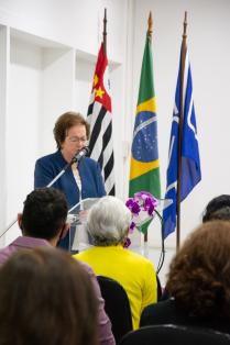 Foto de uma senhora discursando. Ela é branca, tem cabelos curtos e lisos, usa óculos, veste uma blusa branca e um blazer azul. Atrás dela, estão hasteadas três bandeiras. Algumas pessoas estão sentadas assistindo à senhora e de costas para a câmera.