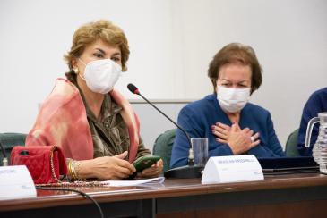 Foto de duas mulheres de máscara sentadas à mesa de madeira. À esquerda, está uma mulher branca falando próxima a um microfone. Ela é branca, tem cabelos curtos e claros, tem olhos escuros e veste uma blusa marrom e um xale rosa. À direita, está uma senhora branca, de cabelos curtos e vestida azul. Ela cruza as mãos em frente ao corpo em sinal de agradecimento. Ao fundo, está uma parede branca.