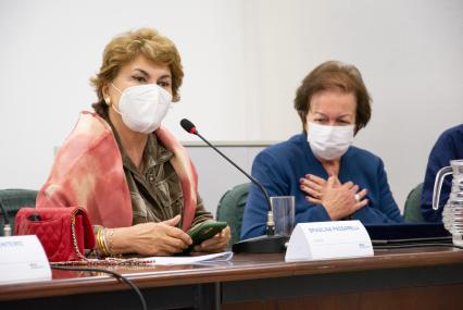 Foto de duas sentadas e usando máscara. À esquerda, está Brasilina, que é branca, tem cabelos curtos, lisos e loiros, veste uma camisa verde e um xale rosa. Ela fala em um microfone. À direita, Margarida, que é branca, tem cabelos curtos, lisos e escuros, veste um terno azul. Ela leva as duas mãos ao coração.