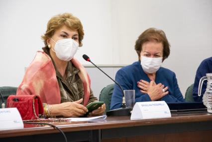 Foto de duas sentadas e usando máscara. À esquerda, está Brasilina, que é branca, tem cabelos curtos, lisos e loiros, veste uma camisa verde e um xale rosa. Ela fala em um microfone. À direita, Margarida, que é branca, tem cabelos curtos, lisos e escuros, veste um terno azul. Ela leva as duas mãos ao coração. 