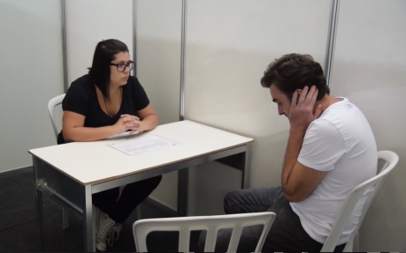 Foto de uma mulher e de um homem sentados próximos a uma mesa. A mulher é branca, tem cabelos lisos e escuros, usa óculos e está vestida de preto. O homem é branco, tem cabelos curtos escuros e veste uma camiseta branca. Ela está apoiada na mesa olhando para o rapaz. Ele está olhando para baixo com as mãos na cabeça e os braços juntos ao corpo.