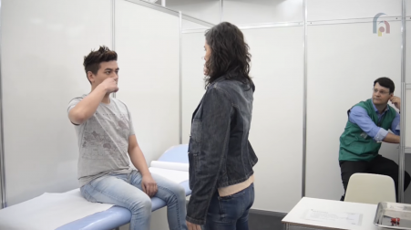 Foto de um atendimento simulado. Um homem branco de cabelos curtos e escuros está sentado em uma maca e apoia um dedo em seu próprio nariz. Uma mulher está em pé na sua frente, ela é branca e tem cabelos longos e escuros. Ao fundo da sala, um homem sentado em um banco assiste à cena, ele é branco e tem cabelos curtos e escuros.