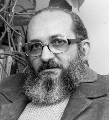 Fotografia em preto e branco de Paulo Freire, homem calvo com barba grisalha e óculos de armação quadrada. Ele olha para a câmera e usa um blazer de veludo e blusa de tricô. Ao fundo é possível ver alguns livros e folhagens.  