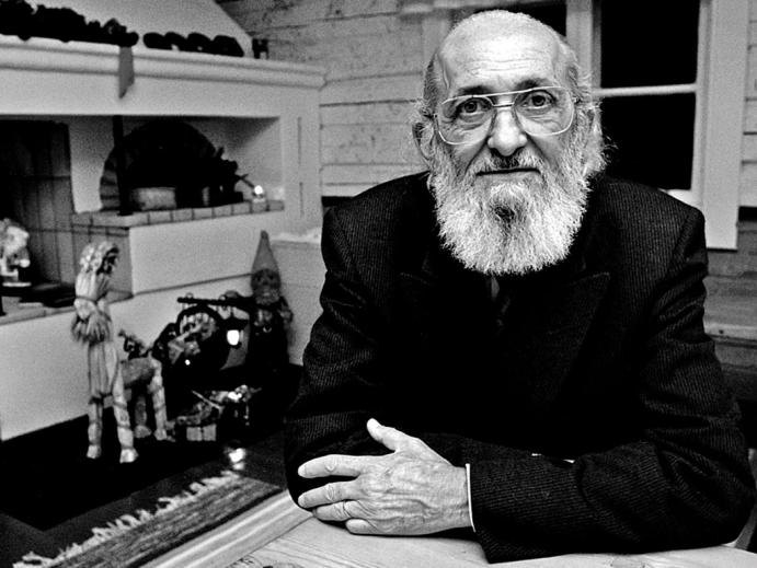 Foto em preto e branco. Paulo Freire, um senhor branco, calvo e de barba comprida e branca, usa óculos com aro de metal em formato quadrado e olha para a câmera enquanto permanece de braços cruzados sobre uma mesa de madeira. Ele usa um terno preto. Atrás dele vemos uma janela e algumas esculturas e objetos de estilo popular  