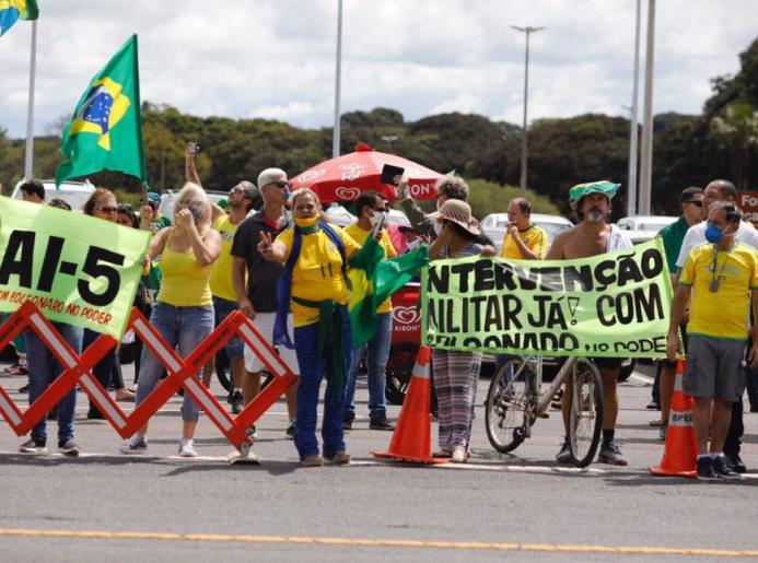 Homens e mulheres, a maioria brancos e de meia idade, vestem roupas verde-amarelas e portam bandeiras do Brasil durante manifestação. Alguns deles mostram cartazes favoráveis ao AI-5 e a intervenção militar com Jair Bolsonaro no poder.