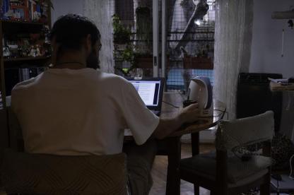 Foto tirada dentro de casa. Um homem, que veste camiseta branca, está de sentado de costas e apoia-se numa mesa. Ele segura uma caneca e encara um notebook. Ao fundo, tem-se outros móveis da casa, algumas plantas e uma janela. A foto é bem escura, o que propicia maior destaque para a luz que o computador emite.