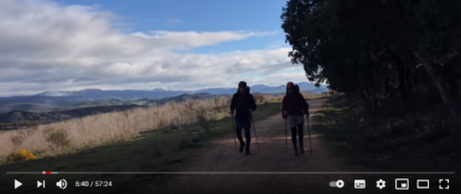 Foto tirada em uma trilha. Duas pessoas aparecem de costas andando com seus equipamentos de peregrinos. A paisagem é composta por um céu com bastante nuvens, montanhas e árvores.