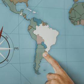 Foto colorida, mão aponta o dedo indicador para países da América Latina em mapa mundi