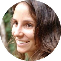 Foto em semi perfil de mulher branca sorrindo. Seus olhos são castanhos e os cabelos longos, ondulados e também castanhos. Ao fundo, uma paisagem verde desfocada.